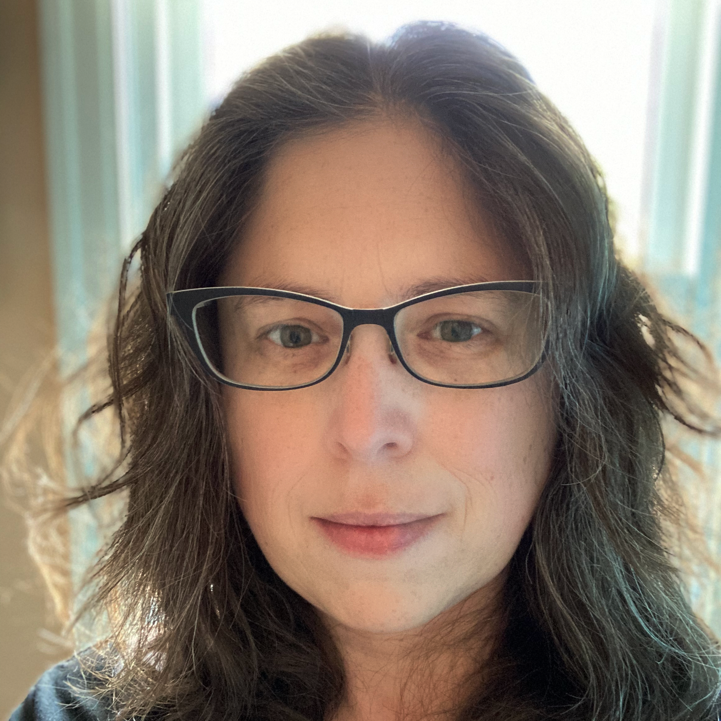 Close-up of Jess in filtered light, with wavy shoulder-length toussled brown hair and wearing black-rimmed glasses, smiling slightly.